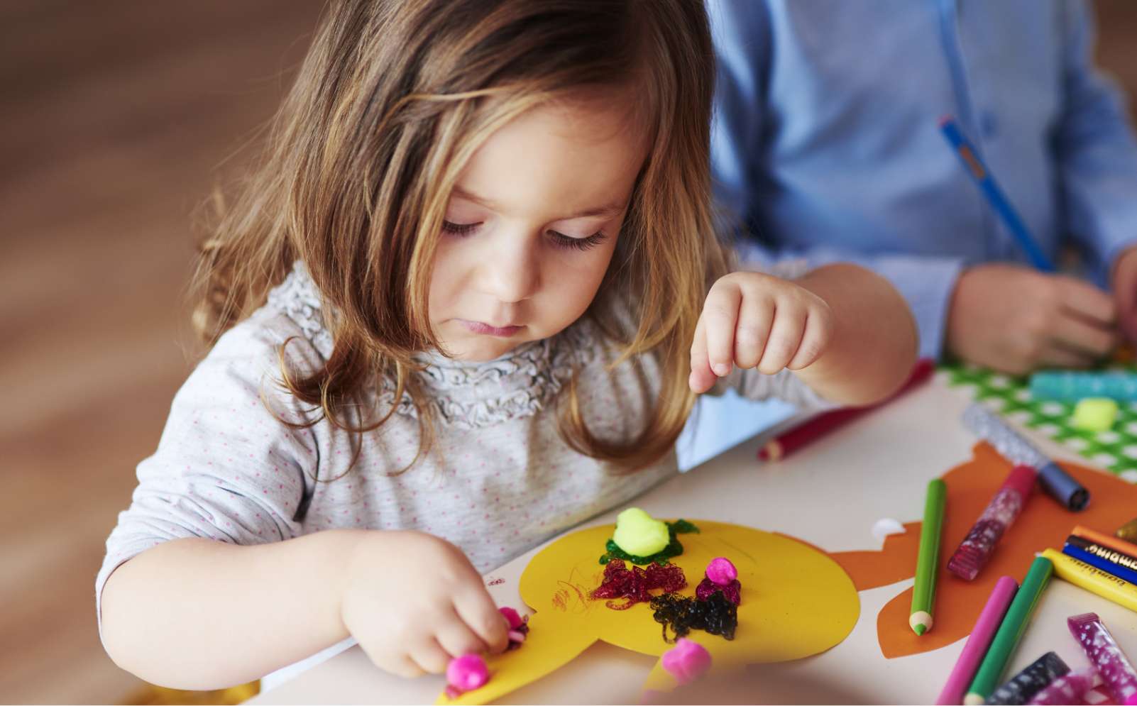 A young girl with blonde hair and wearing a grey t shirt is doing Easter craft, sticking shapes to a yellow paper rabbit.