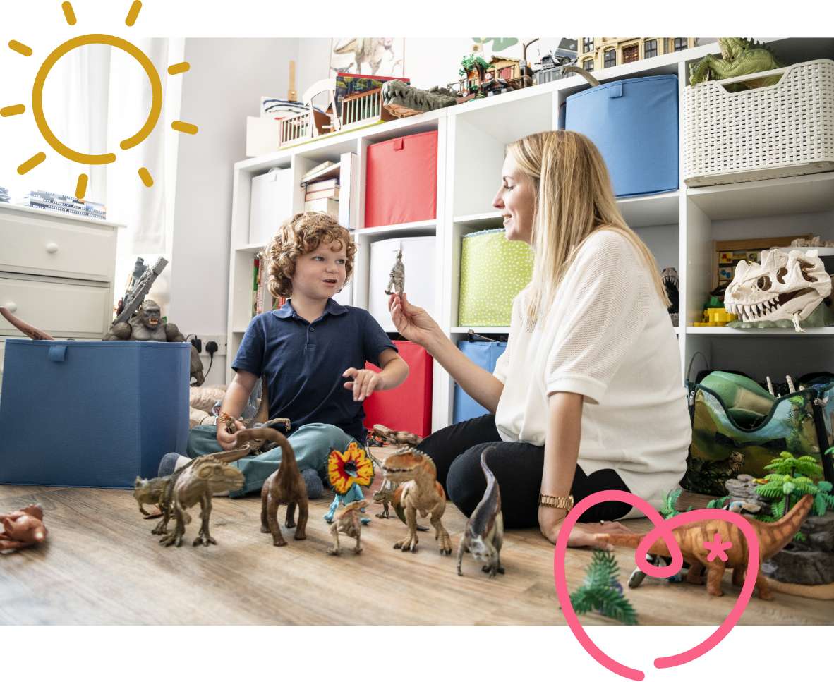A lady in a white top is sat on the floor with a young boy in a blue t shirt. They are playing with plastic dinosaurs.