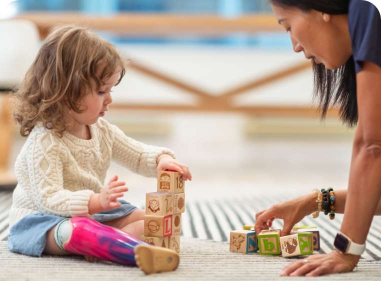 A young girl is sitting on a rug on the floor. She has a prosthetic leg and is playing with alphabet wooden blocks. She is building a tower. Opposite her an Asian woman is sitting on the floor, she is choosing a block from a group in front of her.