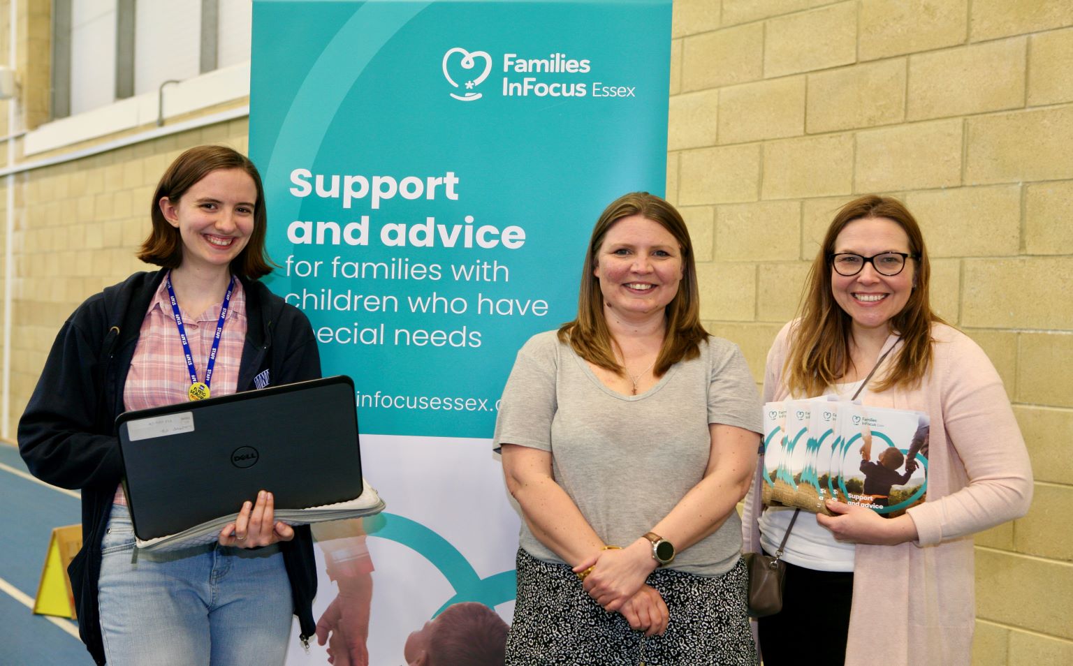 Three Families InFocus staff standing in front of a Families InFocus banner. One is holding a laptop and one is holding leaflets.