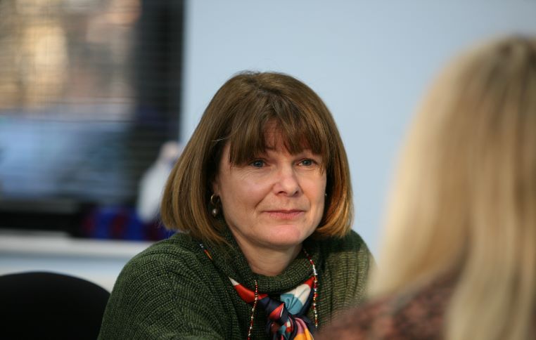 Family Support Advisor Tereasa is sat at her desk, looking at a colleague. She is wearing a green jumper.