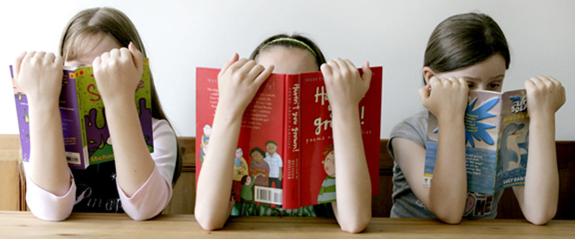 Three girls sitting at a desk holding reading books over their faces