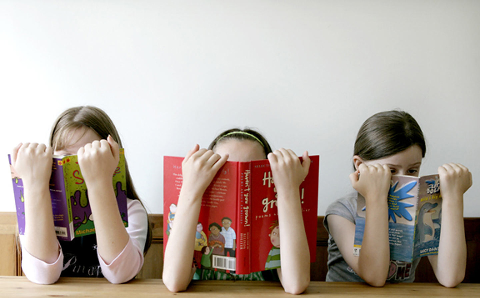 Three girls sitting at a desk holding reading books over their faces