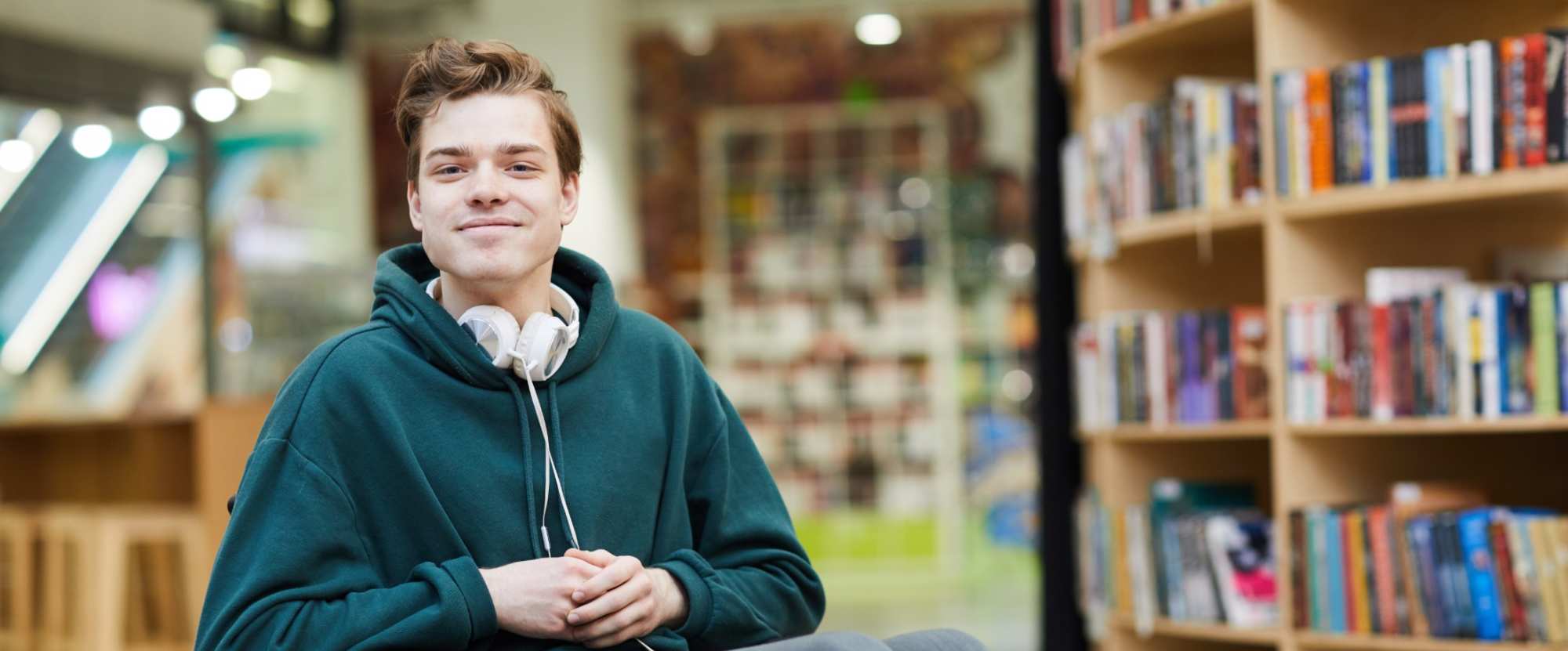 A Young man, wearing a green jumper is sitting in a wheelchair. He is in a library, He is smiling with his arms across his lap.