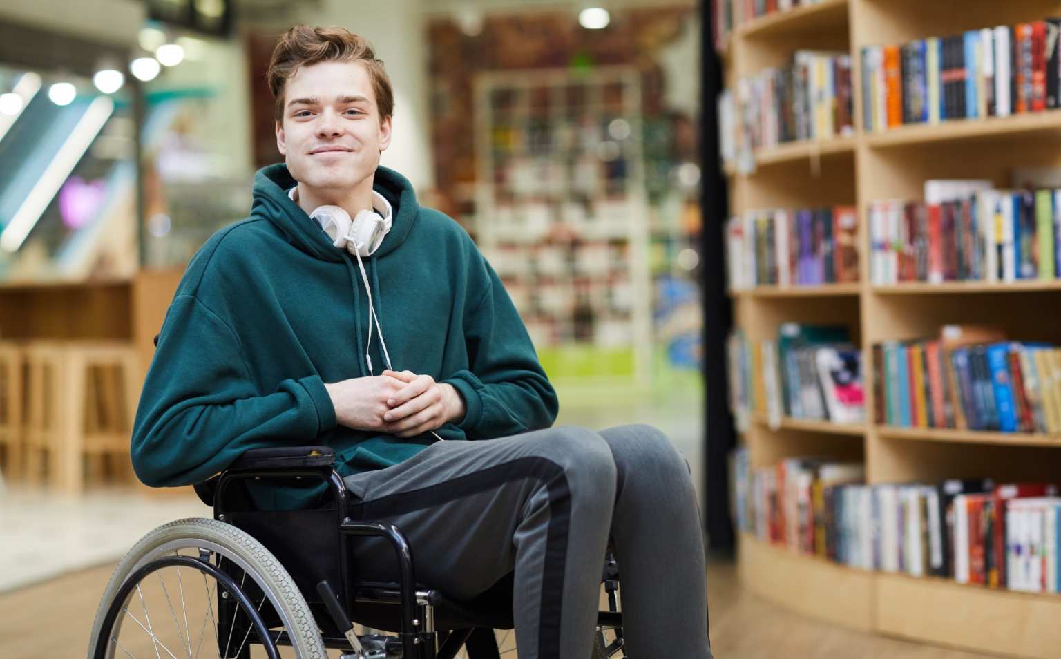 A Young man in a wheelchair is in a library, He is smiling with his arms across his lap.