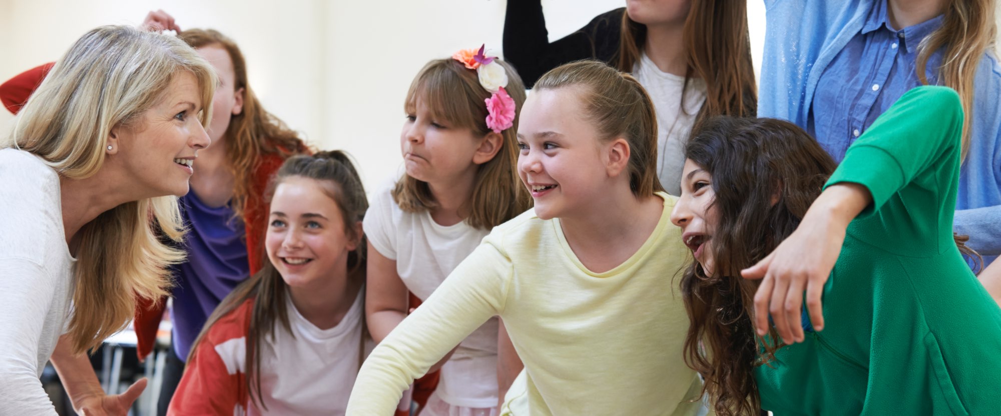 A group of girls are mid way through an acting or movement class. They are wearing brightly colooured tops. They are all looking at an adult who is a teacher, some are happy, some are having fun and one is pulling a silly face.