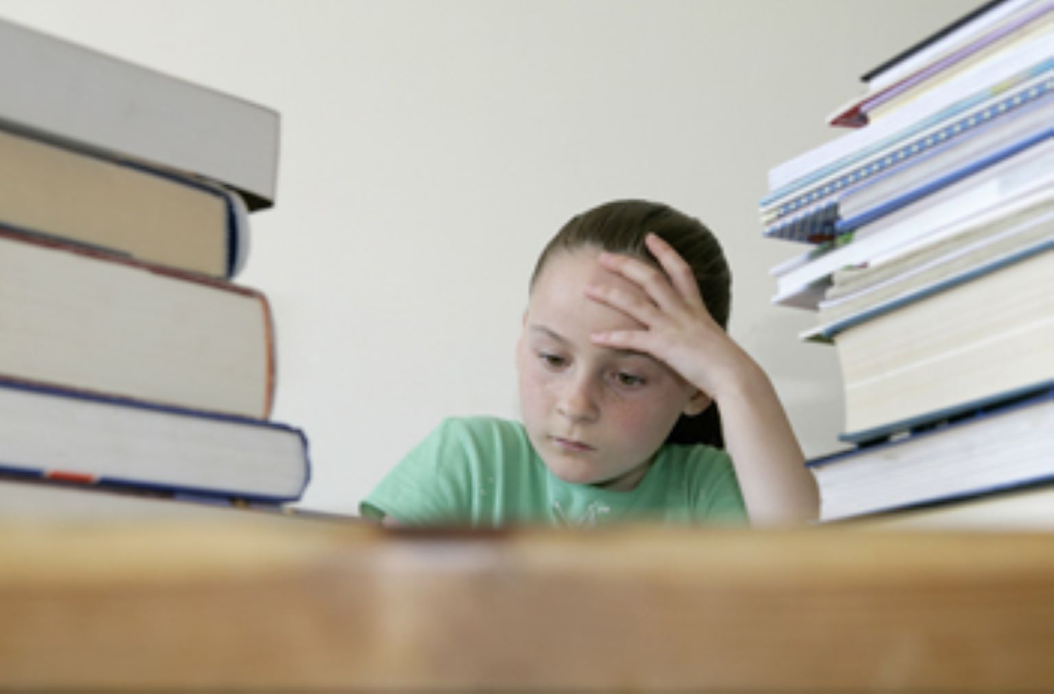 A child is sitting at a table. Her head is resting on her hand, and her arm is bent with her elbow on the table. She is looking down at something on the table. There are two piles of books either side of her. She is concentrating at something on the table in front of her.