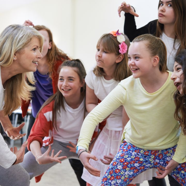 A group of girls are mid way through an acting or movement class. They are wearing brightly coloured tops. They are all looking at an adult who is a teacher, some are happy, some are having fun and one is pulling a silly face.
