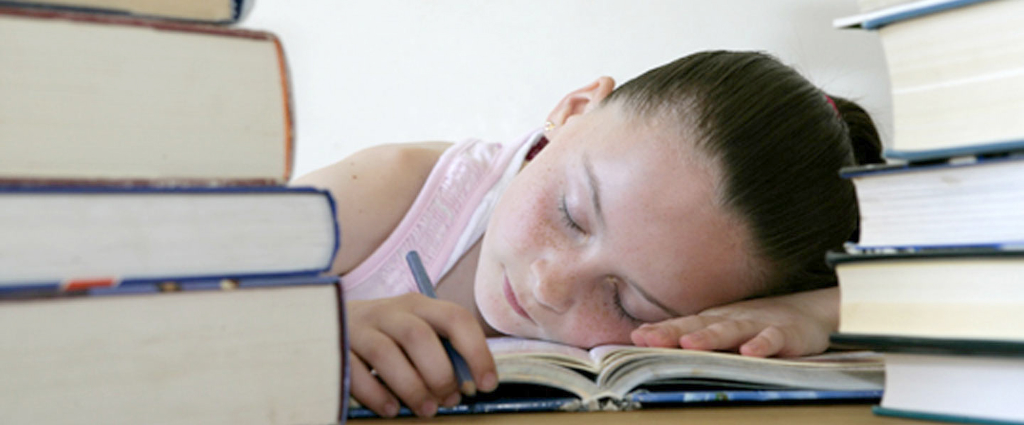 A child is sitting at a table, asleep with her head resting on a book. On the table, either side of her are two piles of books.