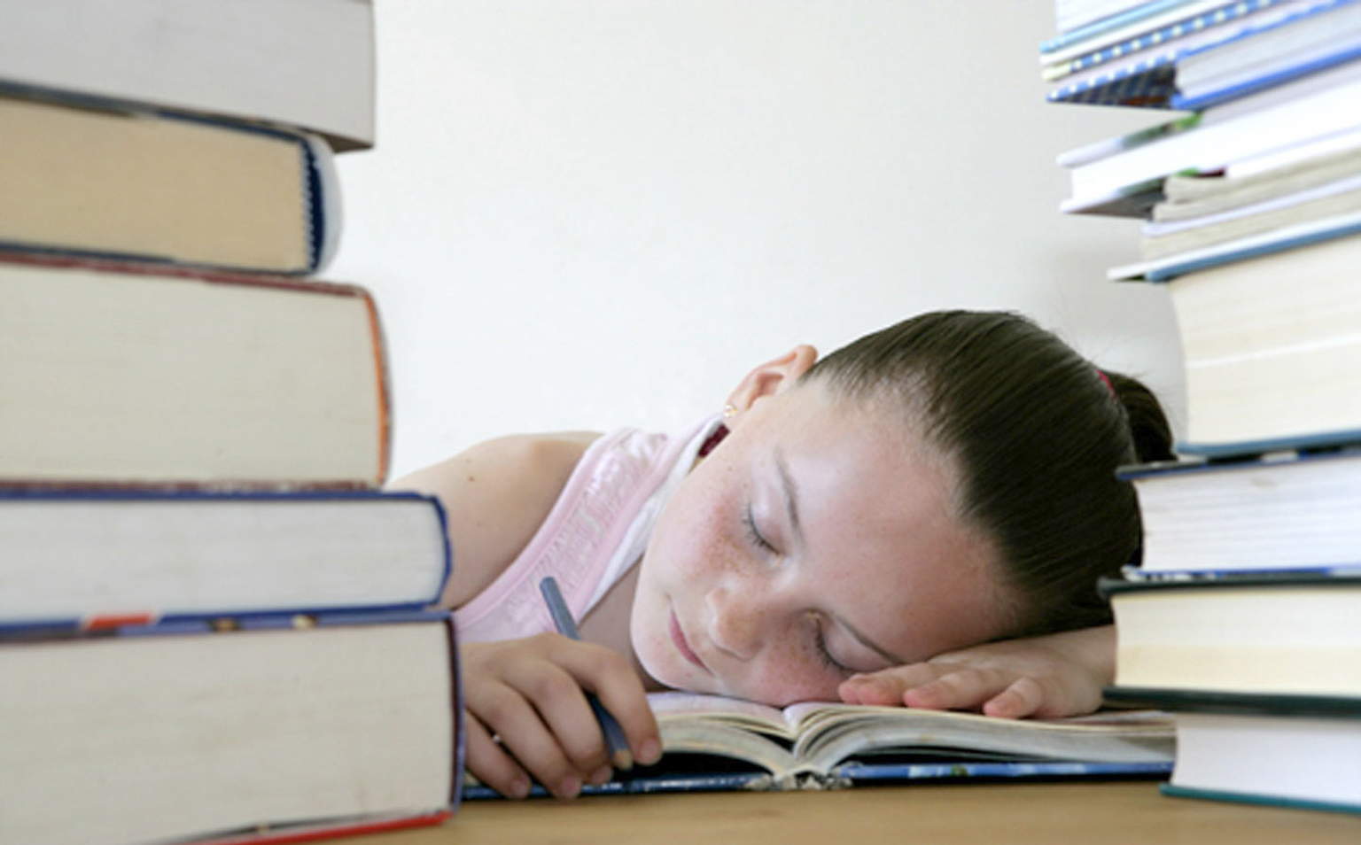 A child is sitting at a table, asleep with her head resting on a book. On the table, either side of her are two piles of books.