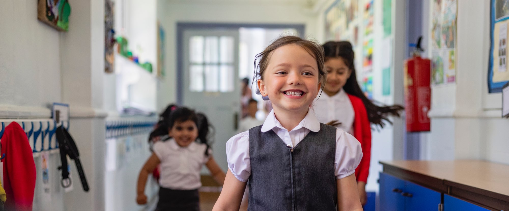 Primary school children in a school corridor, smiling. The girl in front is wearing a white shirt and a grey dress.