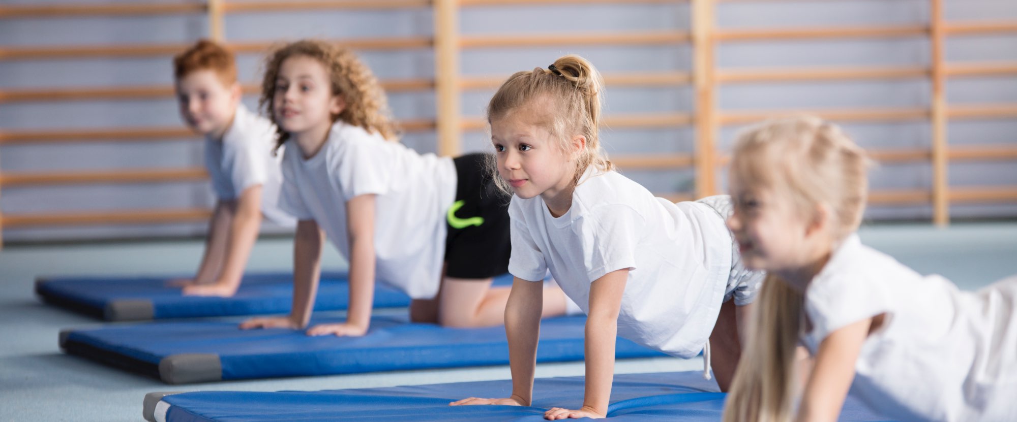 Children doing yoga in their PE lesson. They are wearing white t shirts and shorts, and are exercising on blue mats.