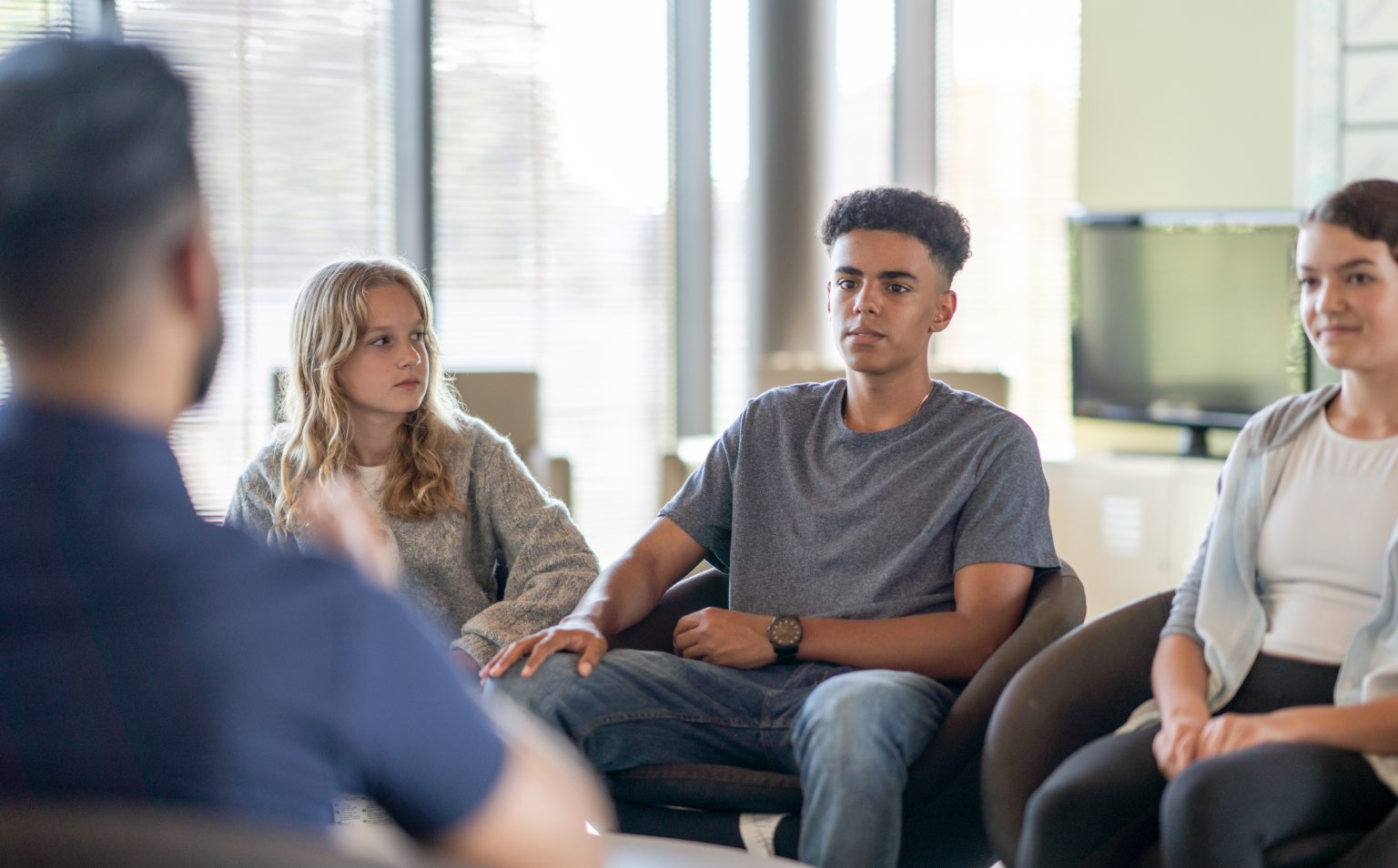 Three young adults are sat in chairs talking to an adult. The girl on the left is blonde and wearing a grey cardigan, the boy in the middle is black and wearing a grey t shirt.