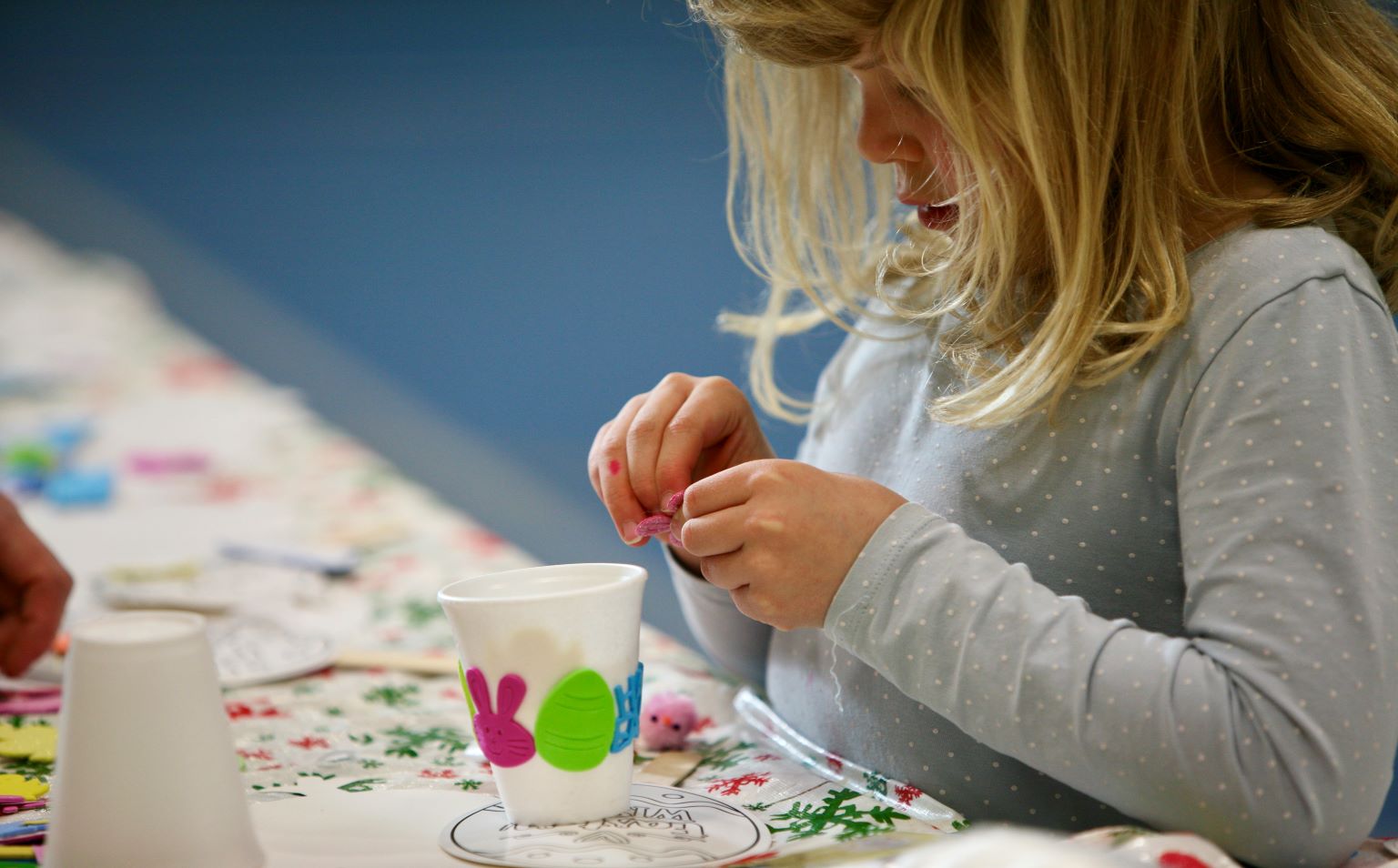 A young girl with long blonde hair is decorating a polystyrene cup with Easter stickers.