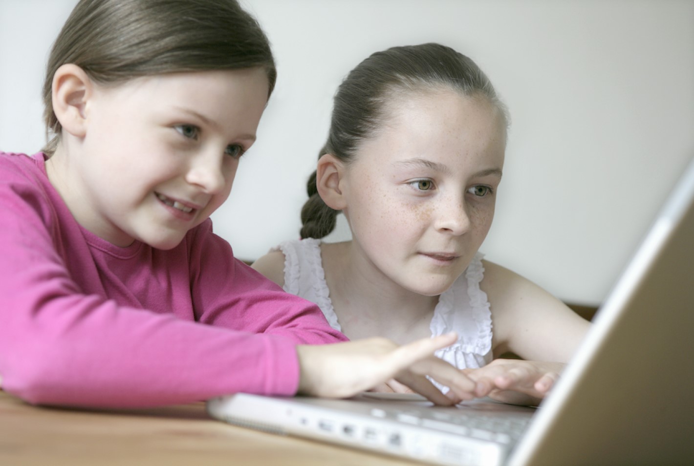 Two girls looking at a laptop and smiling. One is wearing a pink to shirt, and the other is wearing a white t shirt.