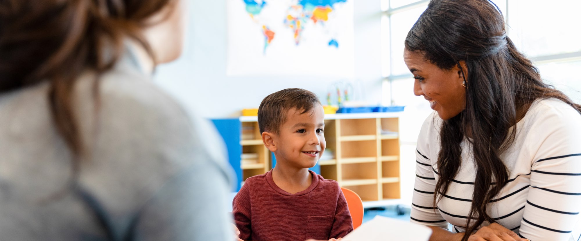 A male child is sitting on a chair in a classroom with two adults. One of the adults has their back to the camera, the other adult female is looking at the child smiling. They look happy.