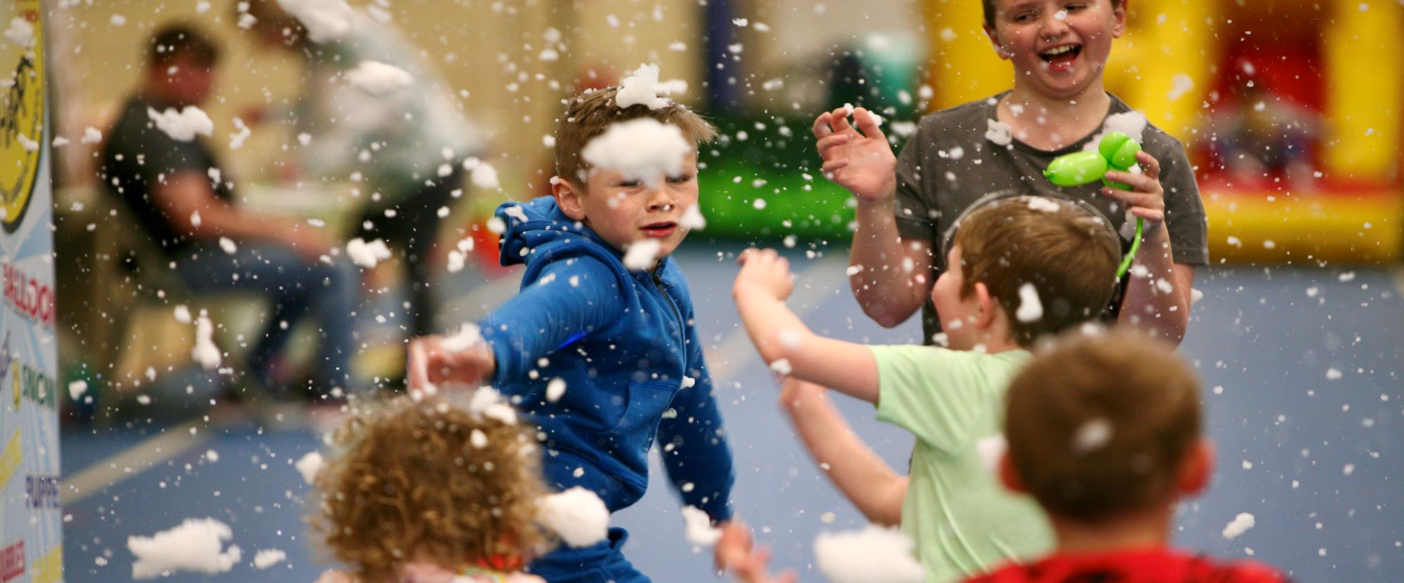 A group of children are at a party at a sports centre. They are trying to catch some fake snow made from a snow machine. They are enjoying themselves.