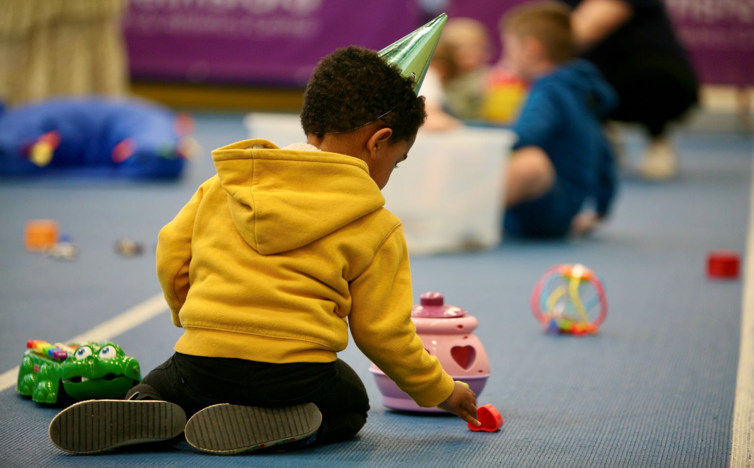 A young black child is sitting on the floor at a party, playing with plastic toys. He is wearing a pointed party hat and a bright yellow hoodie. He is facing away from the camera playing with toys which are on the floor.