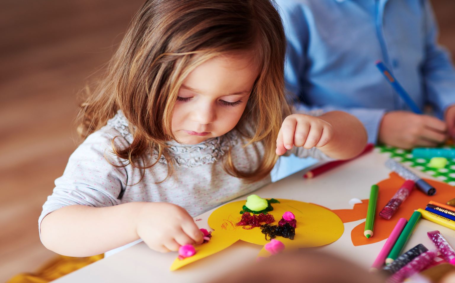 A young girl with blonde hair and wearing a grey t shirt is doing Easter craft, sticking shapes to a yellow paper rabbit.