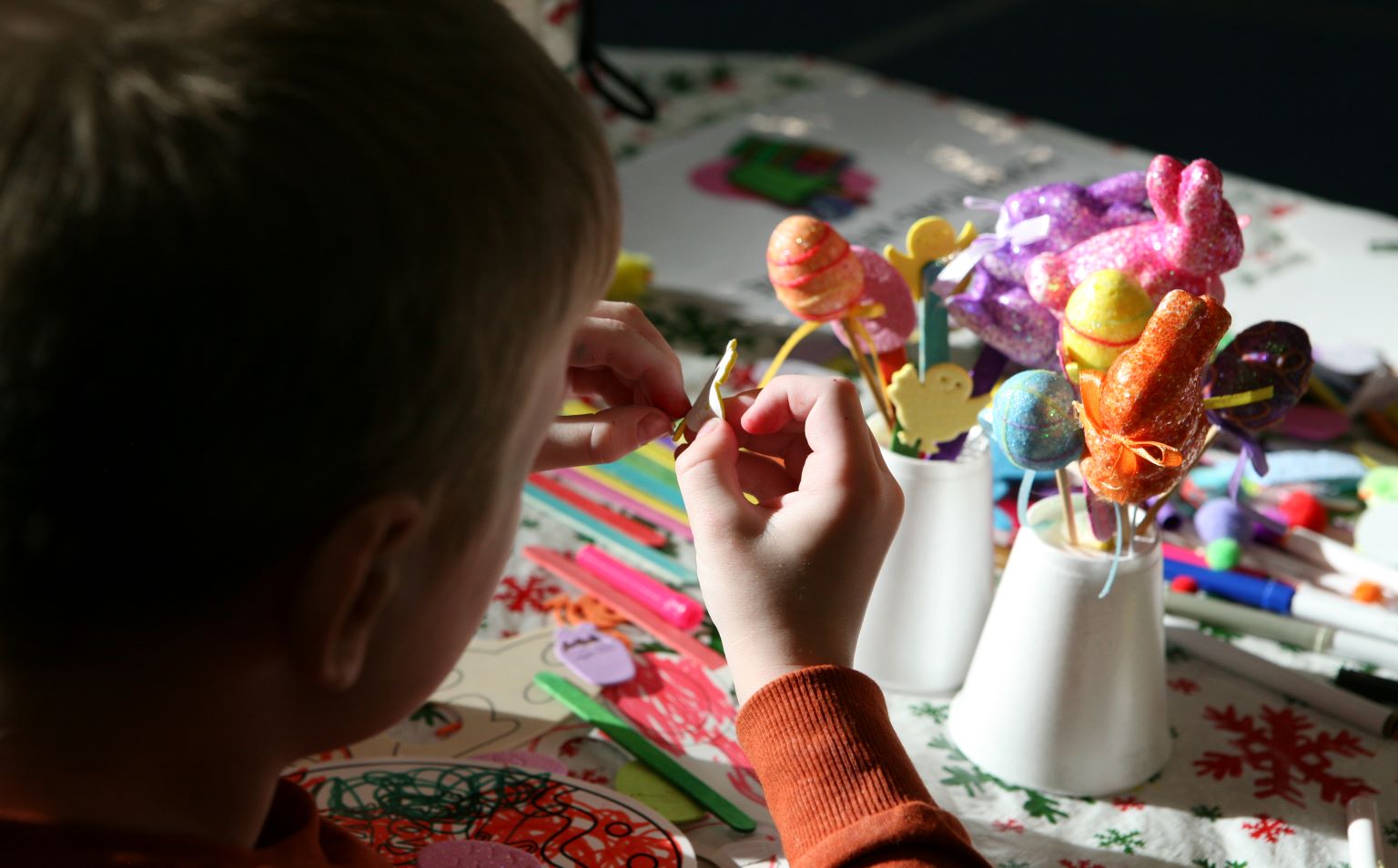 A child is sitting at a table which has easter arts and drafts on it. He is facing away from the camera and he is pealing the back off a sticker to decorate a paper plate.