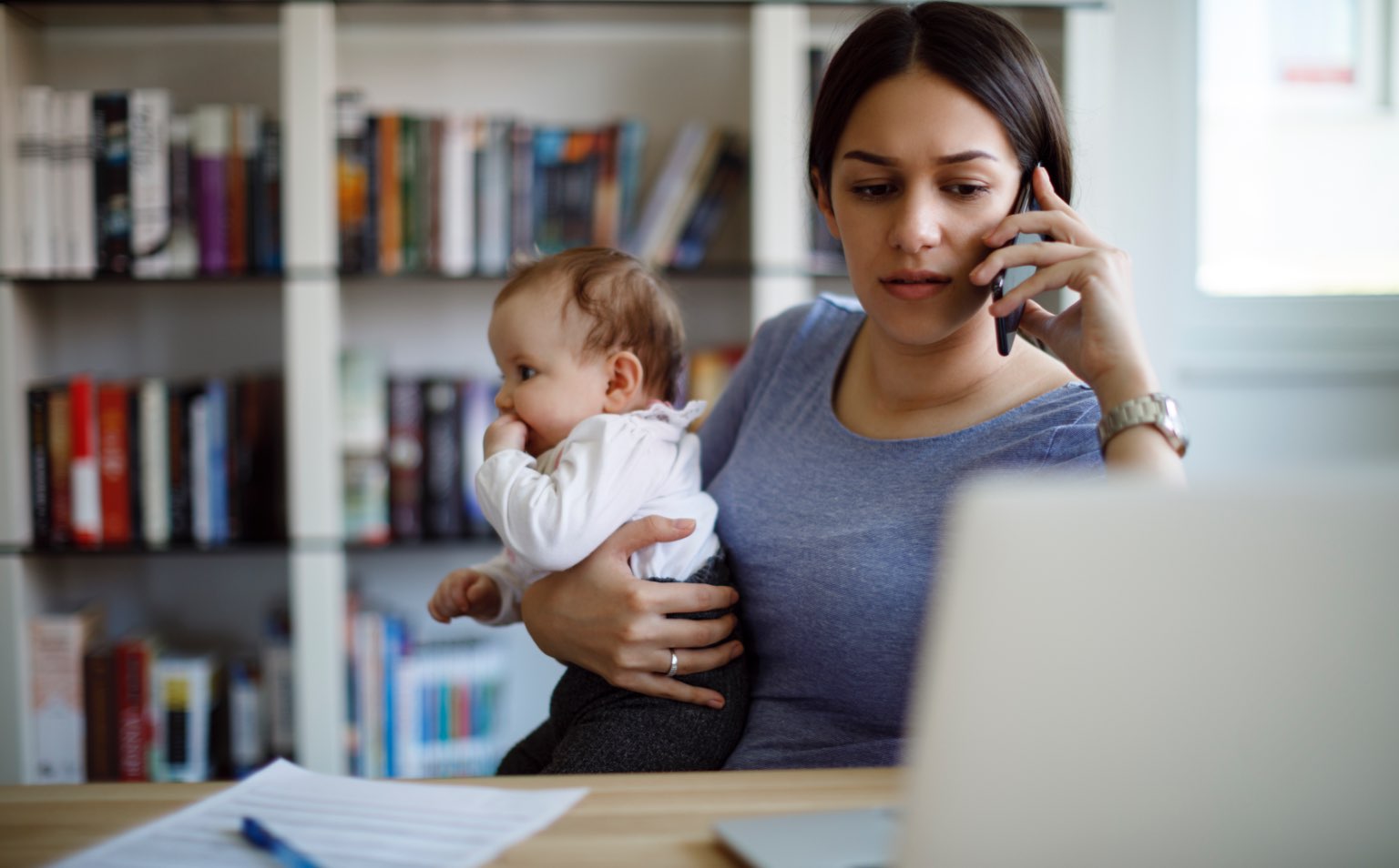 A woman is on phone in front of a laptop holding a baby. She is wearing a blue t shirt and the baby is facing away from her, with his had in his mouth.