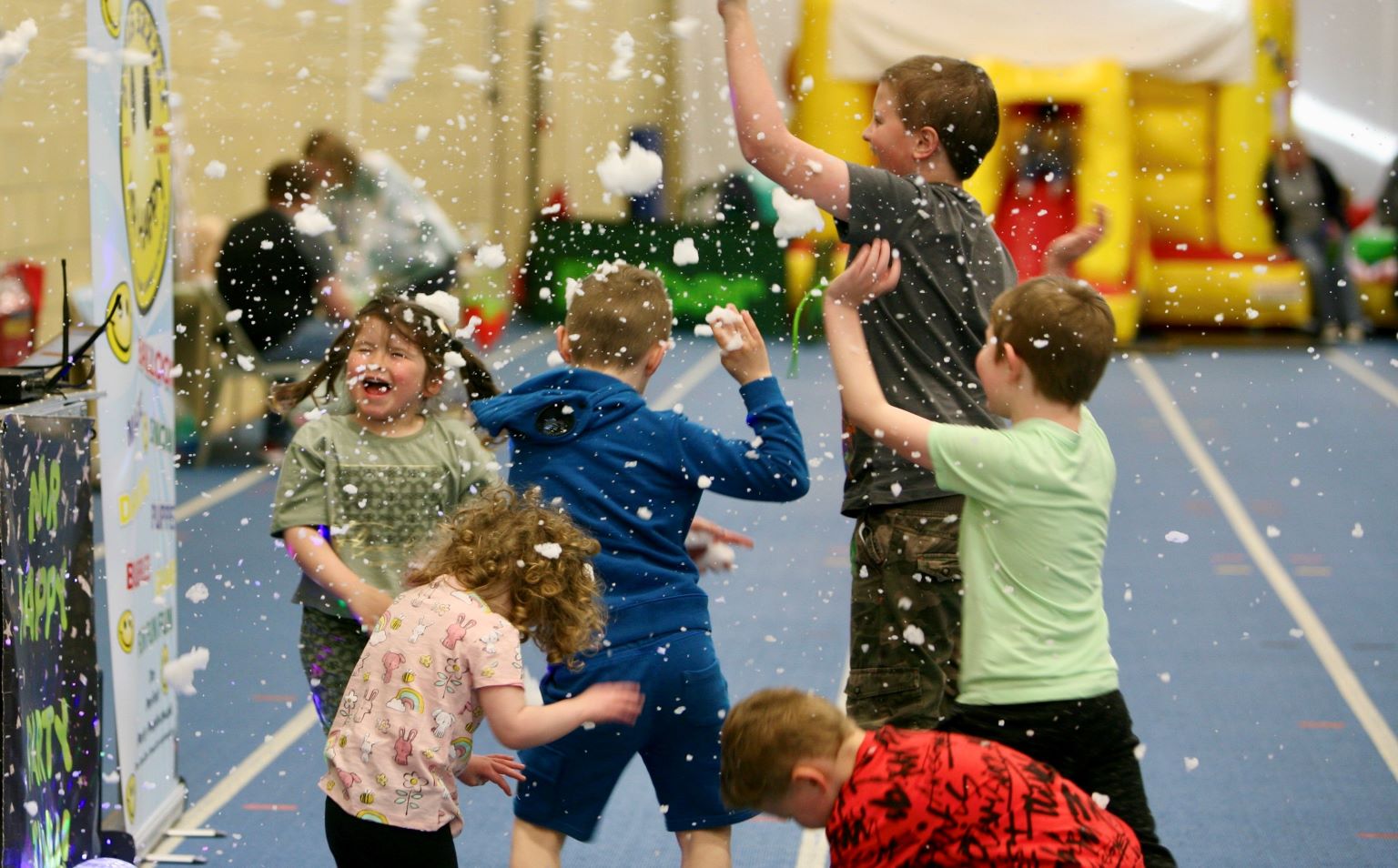 Five children are at a party in a sports centre. In the background there is a bouncy castle. They are laughing and enjoying themselves. Some are jumping up and down and waving their arms. They are catching snow from a snow machine.