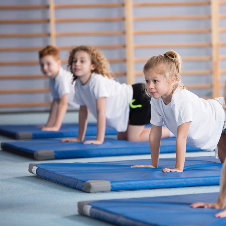 Children doing yoga in their PE lesson. They are wearing white t shirts and shorts, and are exercising on blue mats.