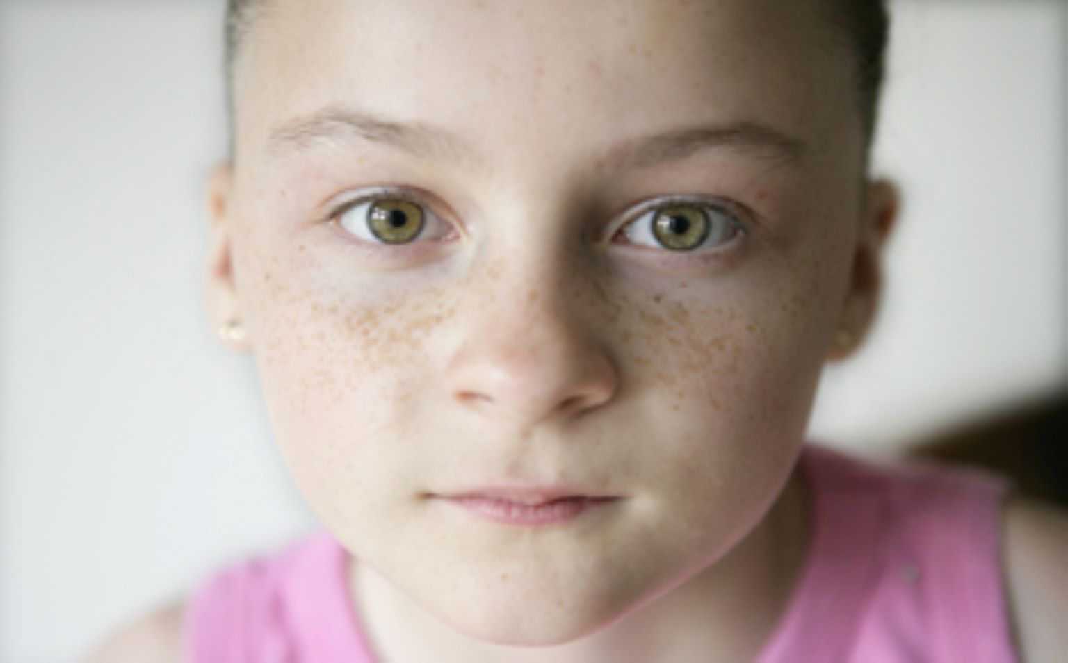 A young girl stares back at the camera. Her hair is pulled back away from her face. She has freckles and green eyes.