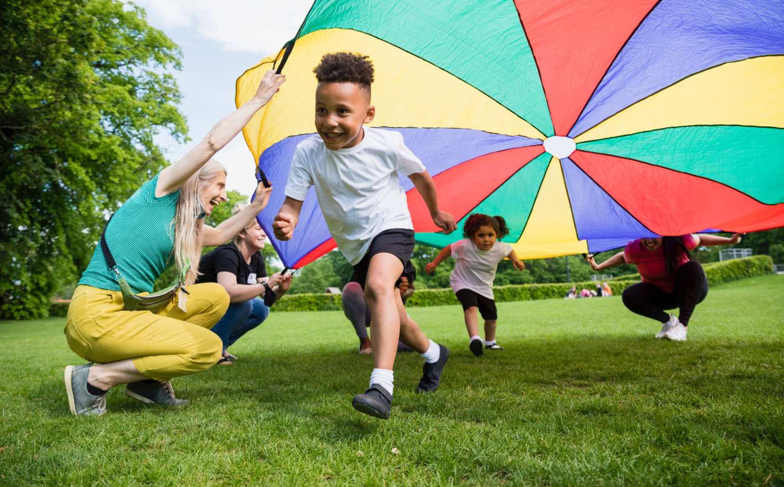Children playing outside with adults, under a colourful parachute.