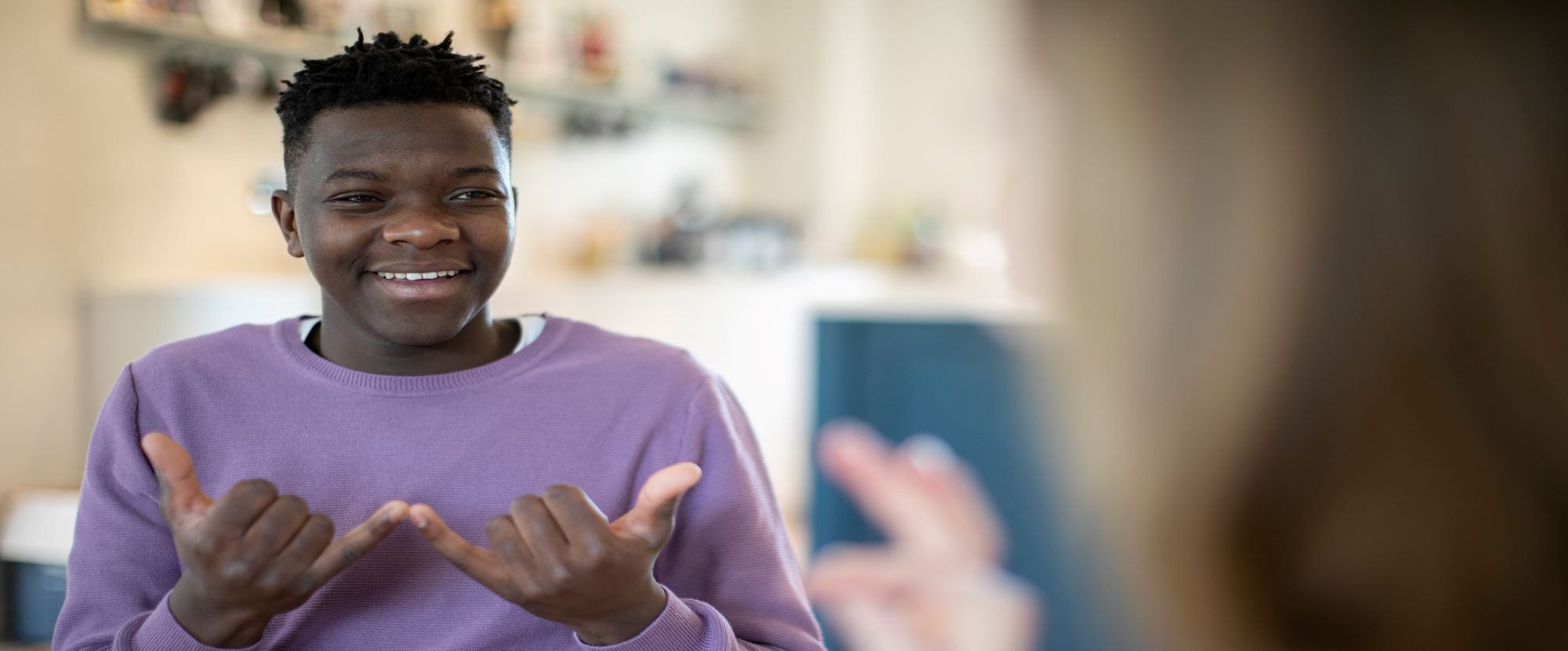A black young man, wearing a purple jumper, using sign language. He is smiling.