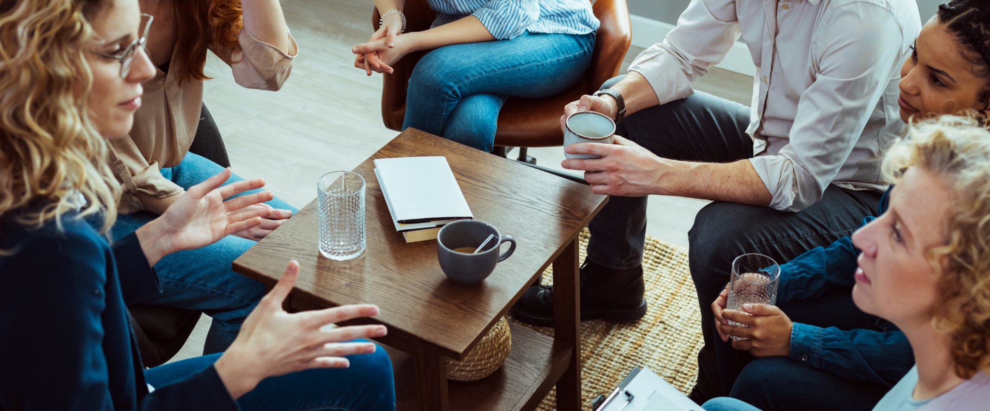 A group of adults are sitting around a coffee table. They are listening to a woman speak. She is gesturing and they are deep in discussion. They look interested in what the woman is saying to them.