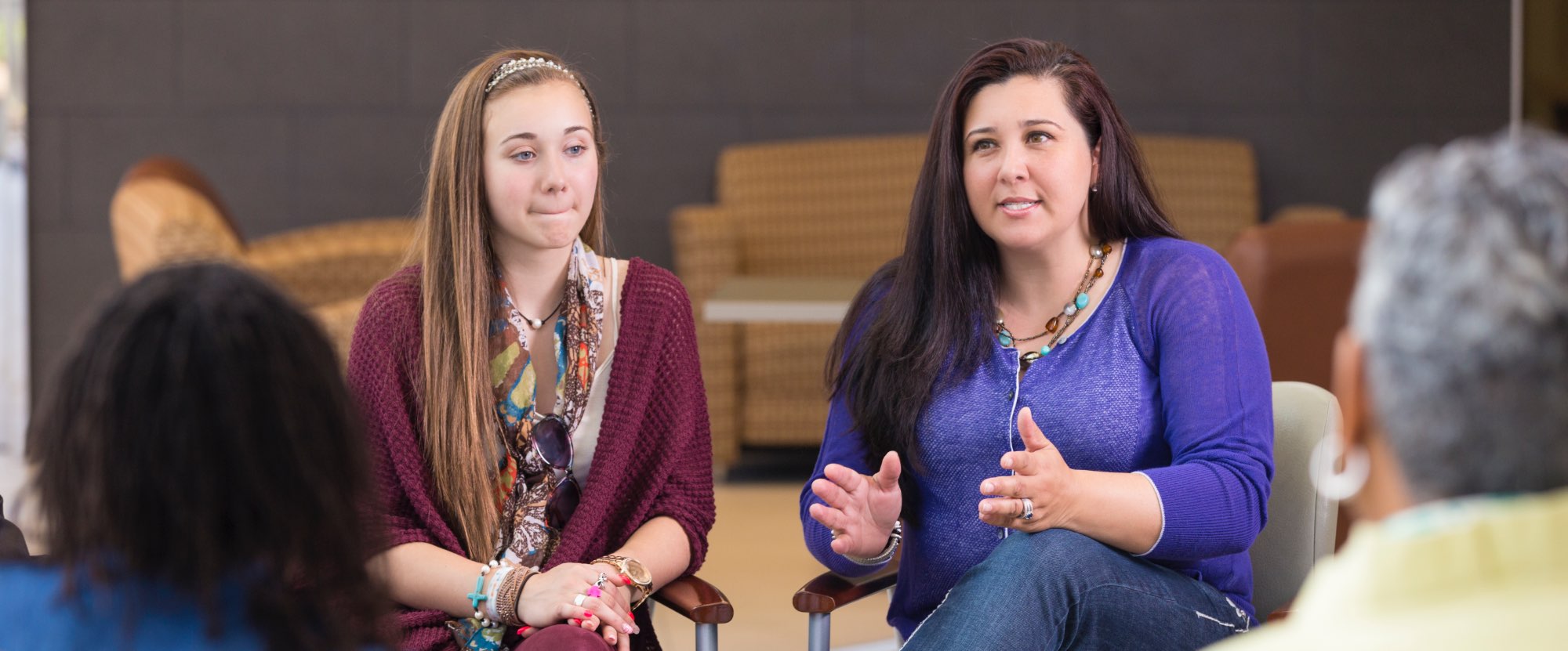 A group of adults are sitting on chairs in a circle. They are in a village hall and are listening to a woman in a purple jumper speak. She is gesturing with her hands and is in deep discussion.