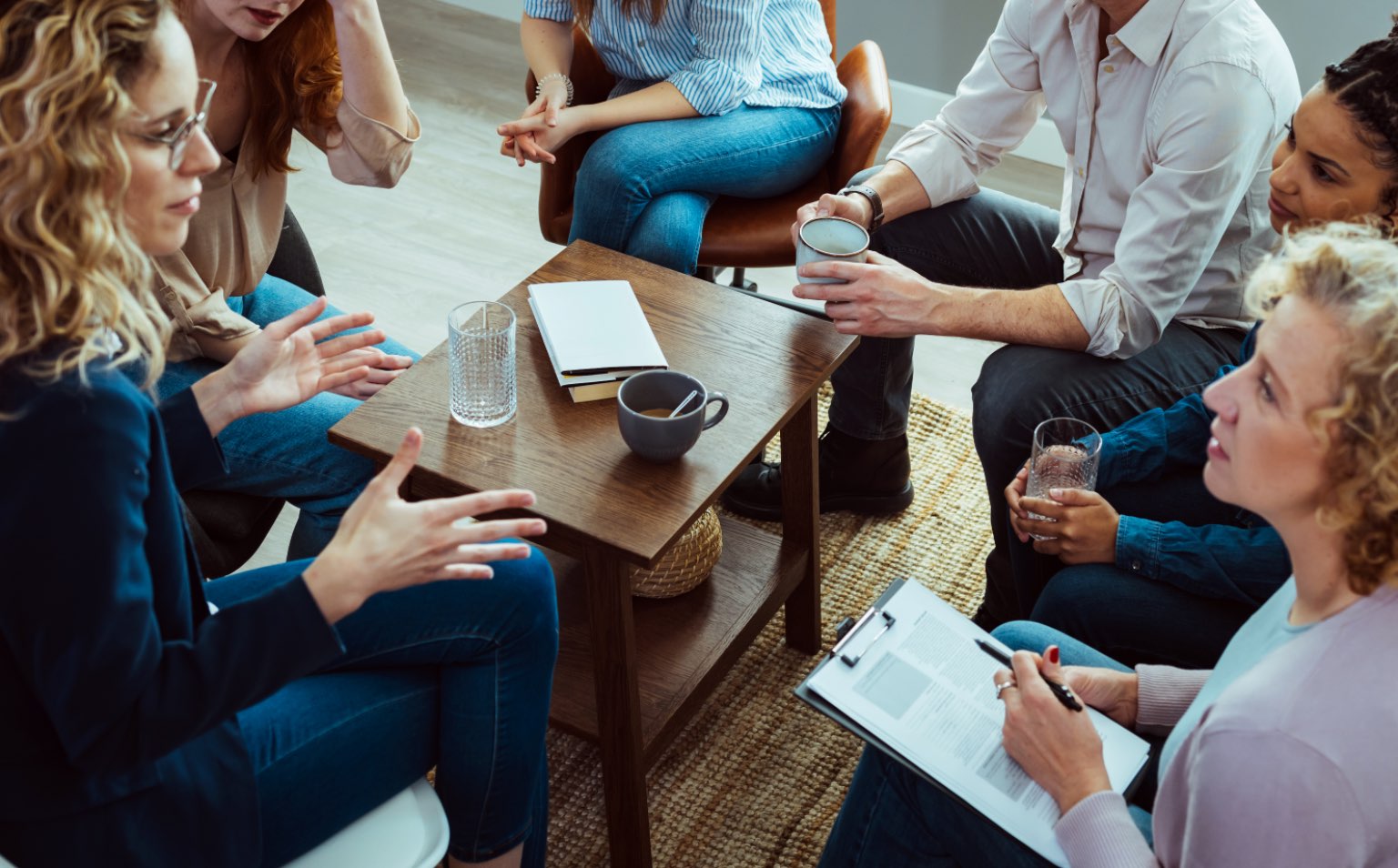 A group of adults are sitting around a coffee table. They are listening to a woman speak. She is gesturing and they are deep in discussion. They look interested in what the woman is saying to them.