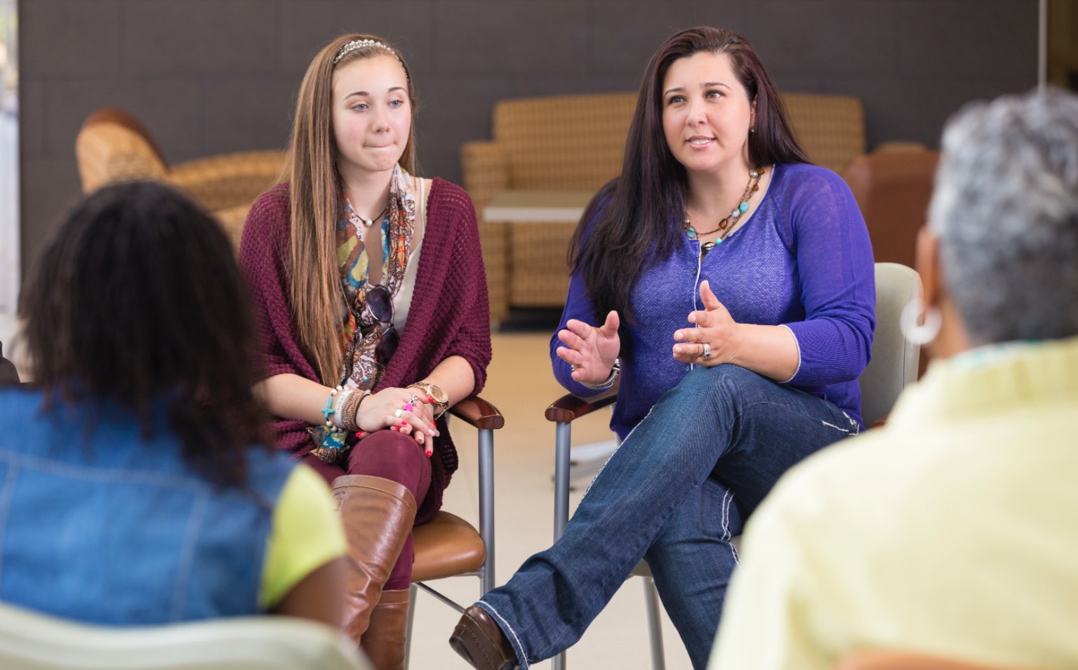 A group of adults are sitting on chairs in a circle. They are in a village hall and are listening to a woman in a purple jumper speak. She is gesturing with her hands and is in deep discussion.